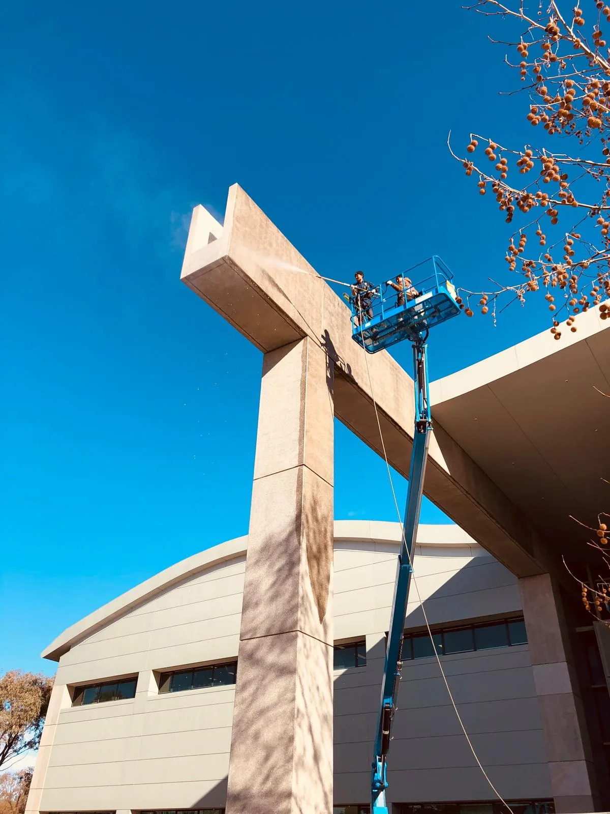 Two workers in a raised boom lift basket pressure washing the top of a tall concrete pylon
