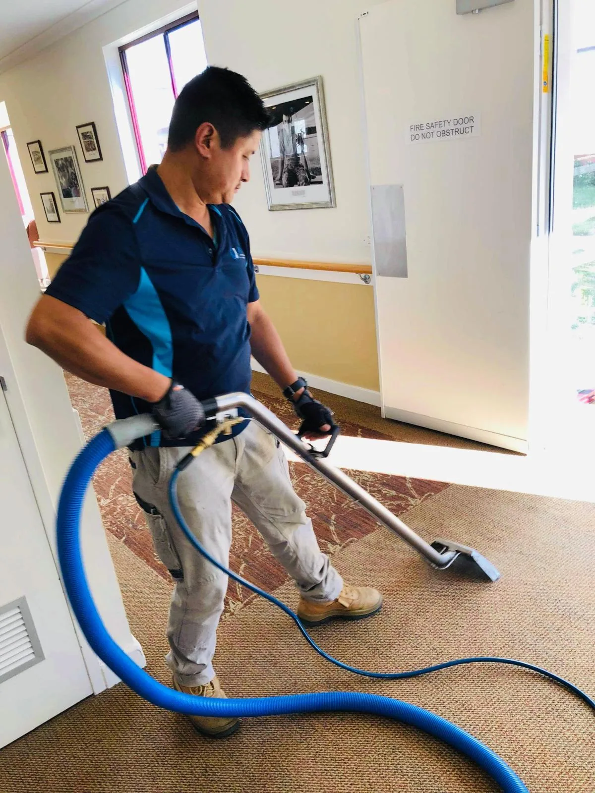 A cleaner working a carpet extraction wand along a corridor carpet, past a wall handrail and a fire door