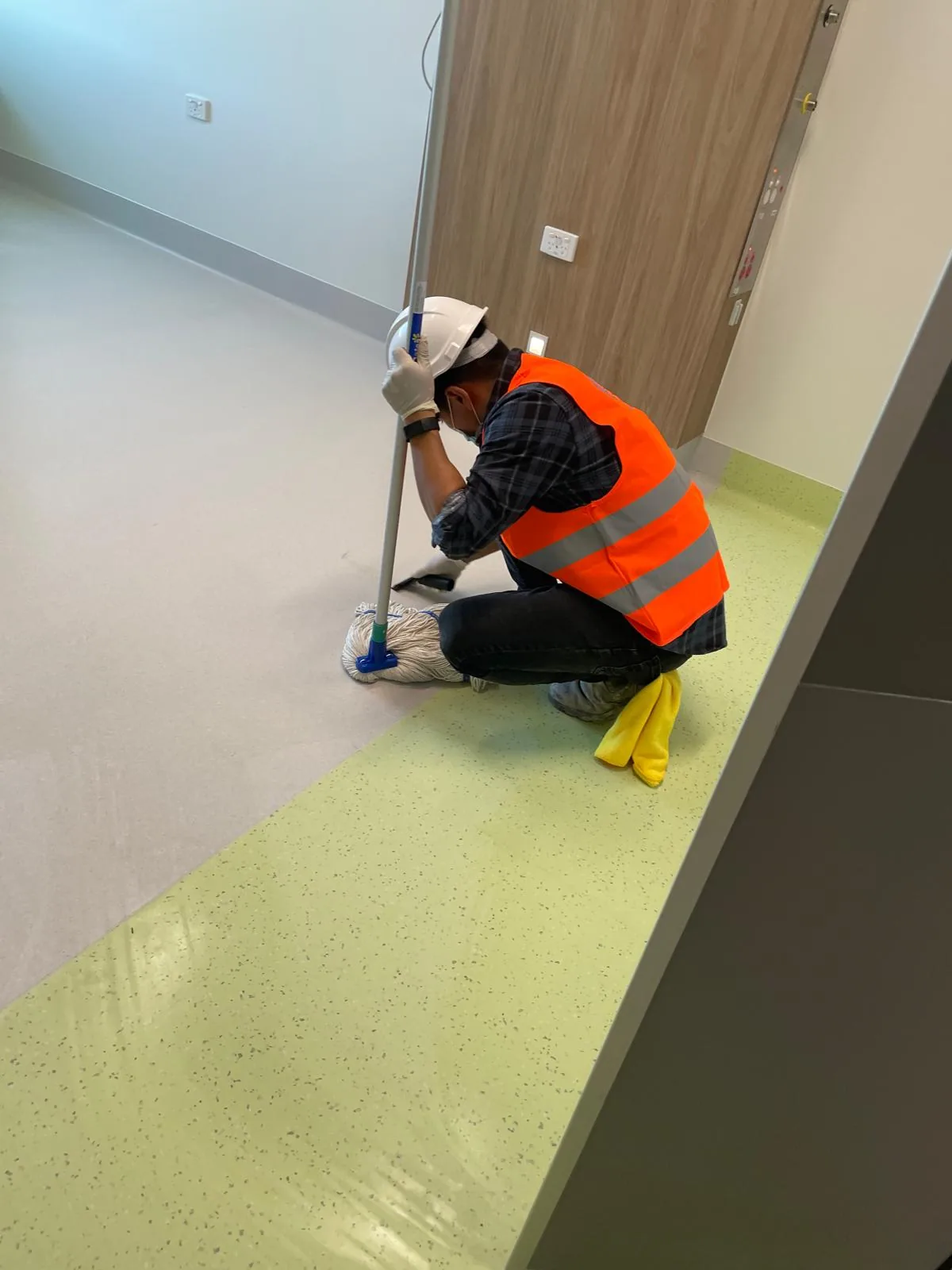 A worker in a hard hat and orange hi-vis kneeling in a hospital ward doorway fitting a mop head