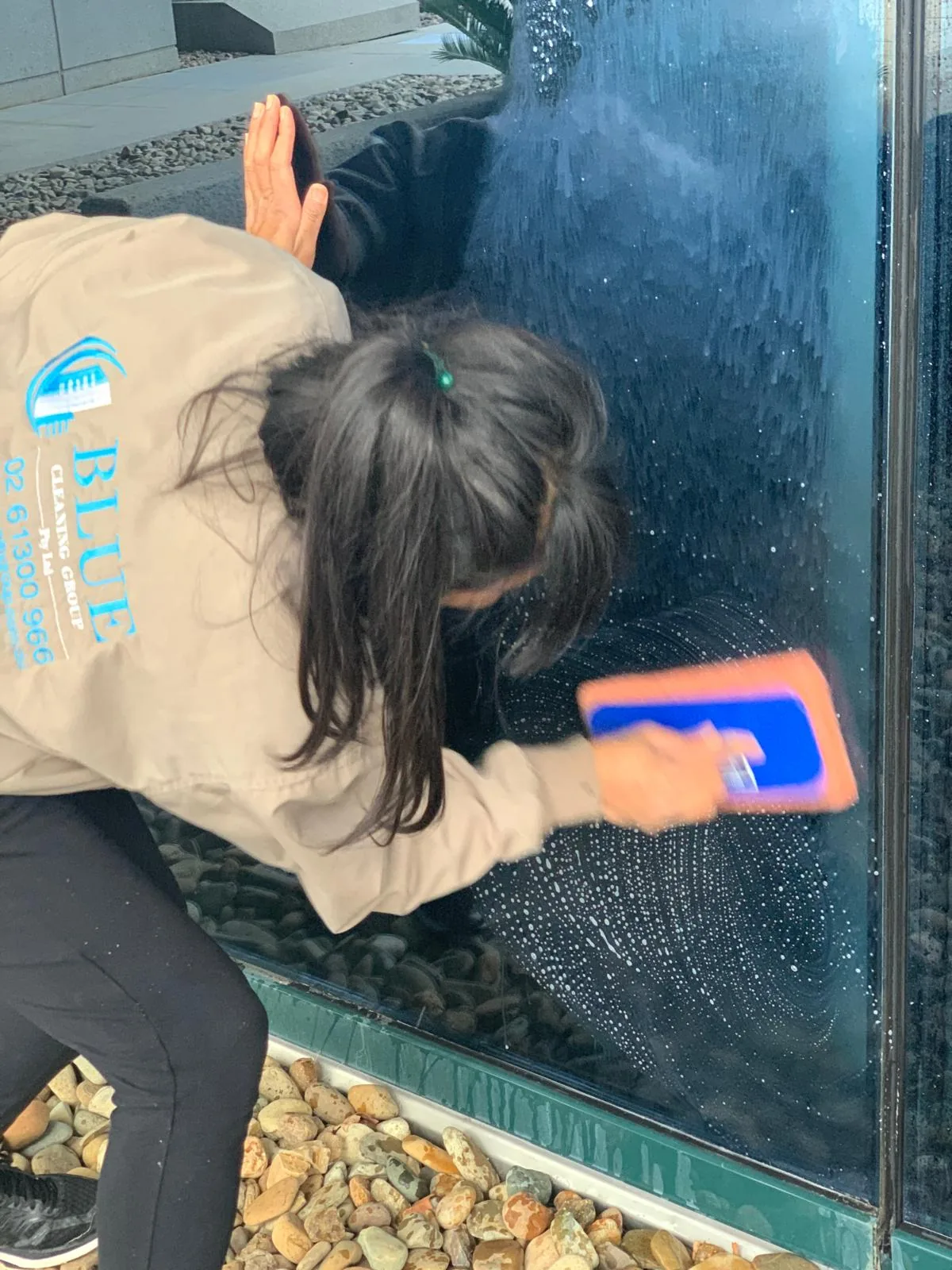 A cleaner in a Blue Cleaning Group jacket crouched over a pebble bed, soaping the lower glass of an exterior panel