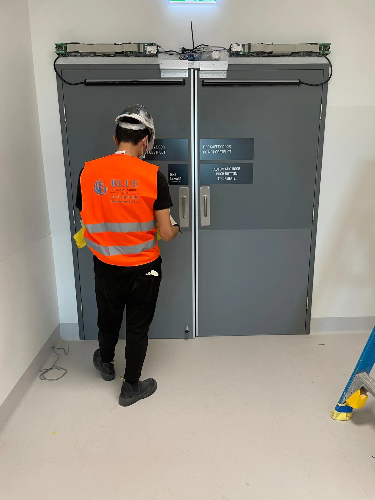 A cleaner in a printed Blue Cleaning Group vest wiping the handle of a grey fire safety door, the door controllers still exposed above