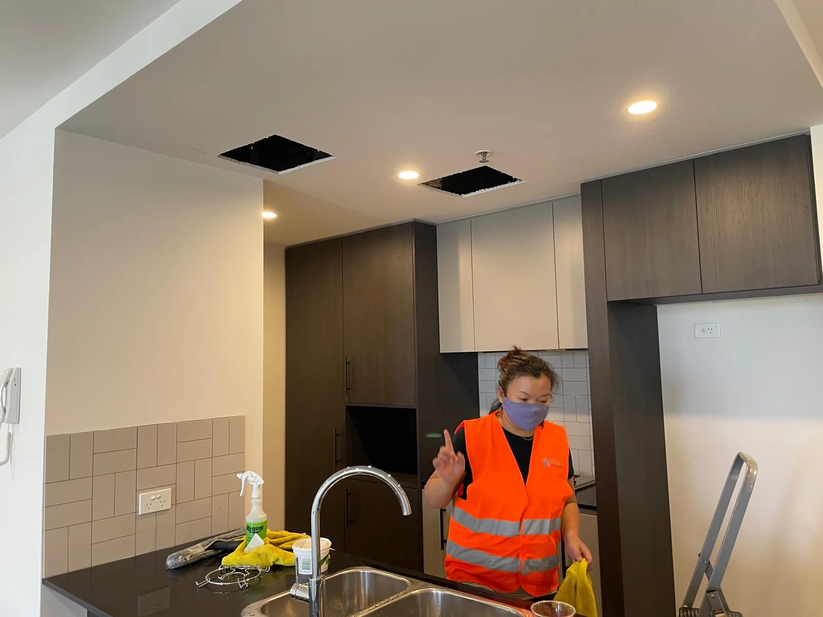 A masked cleaner in orange hi-vis wiping the sink and black stone bench of a new apartment kitchen, ceiling panels still open above