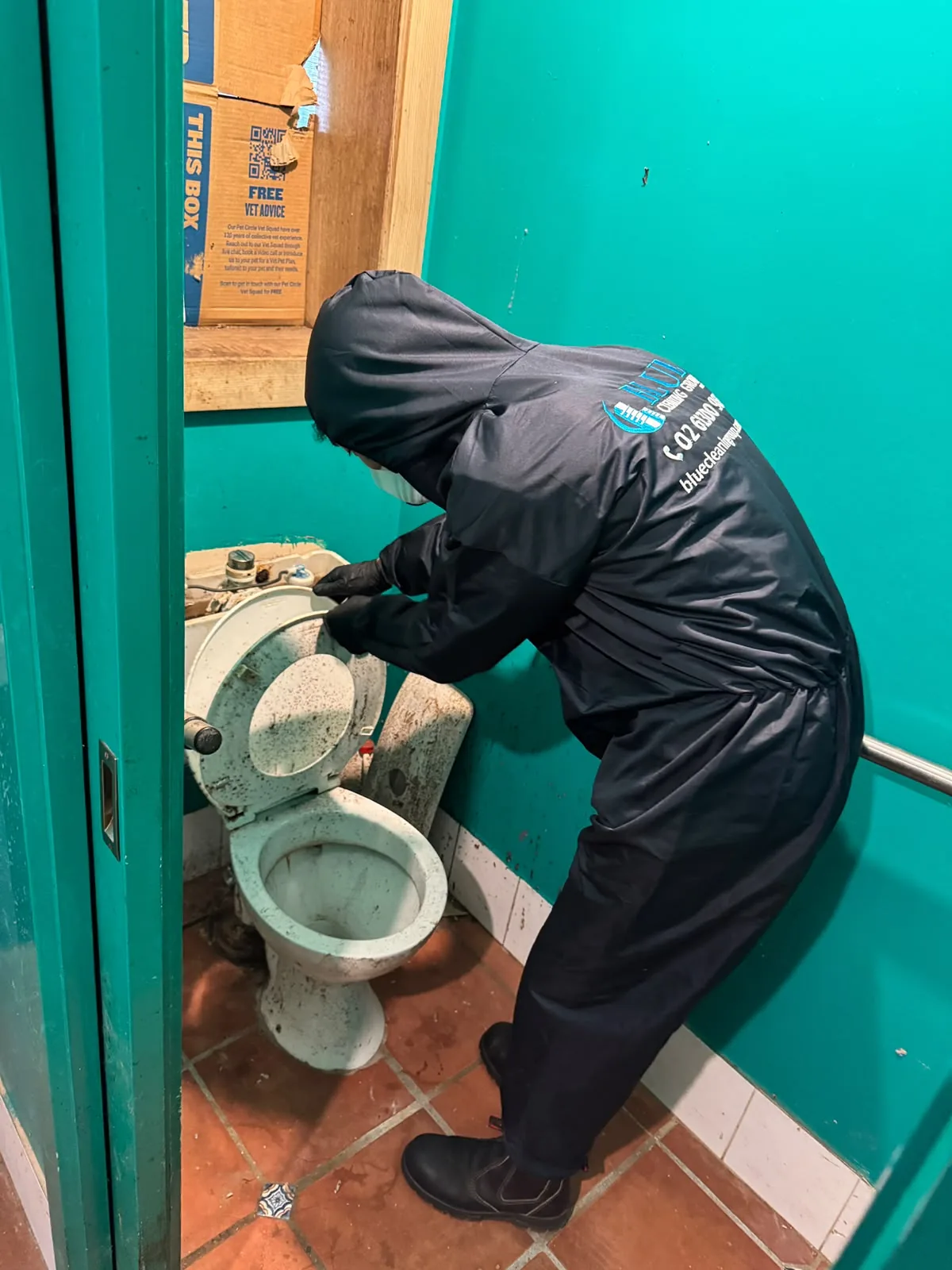 A cleaner in a hooded Blue Cleaning Group coverall beside a heavily soiled toilet in a teal-walled cubicle, before cleaning