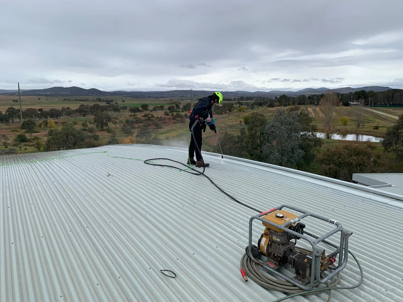 A harnessed operator in a green hard hat washing along a roof ridge with a lance