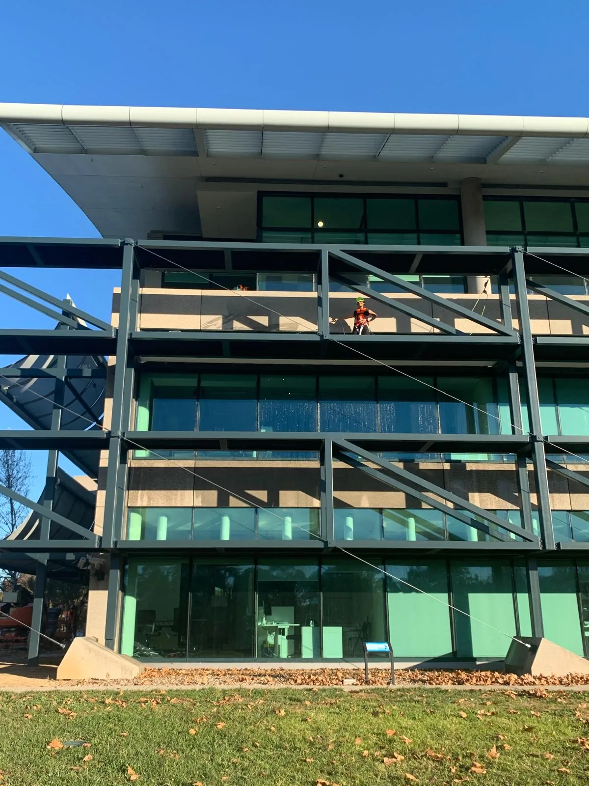 A harnessed worker in orange hi-vis washing a steel-framed glazed facade from a ledge two storeys up