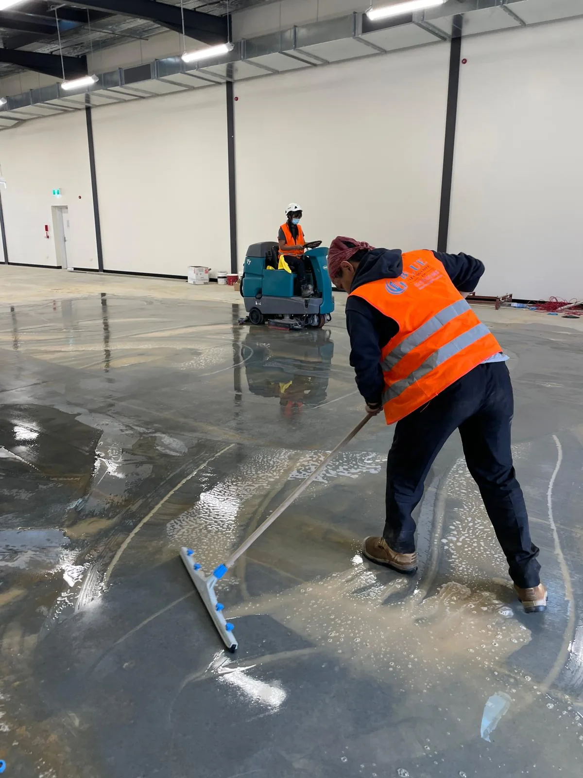 A worker in orange Blue Cleaning hi-vis squeegeeing water across a wet slab in an empty warehouse