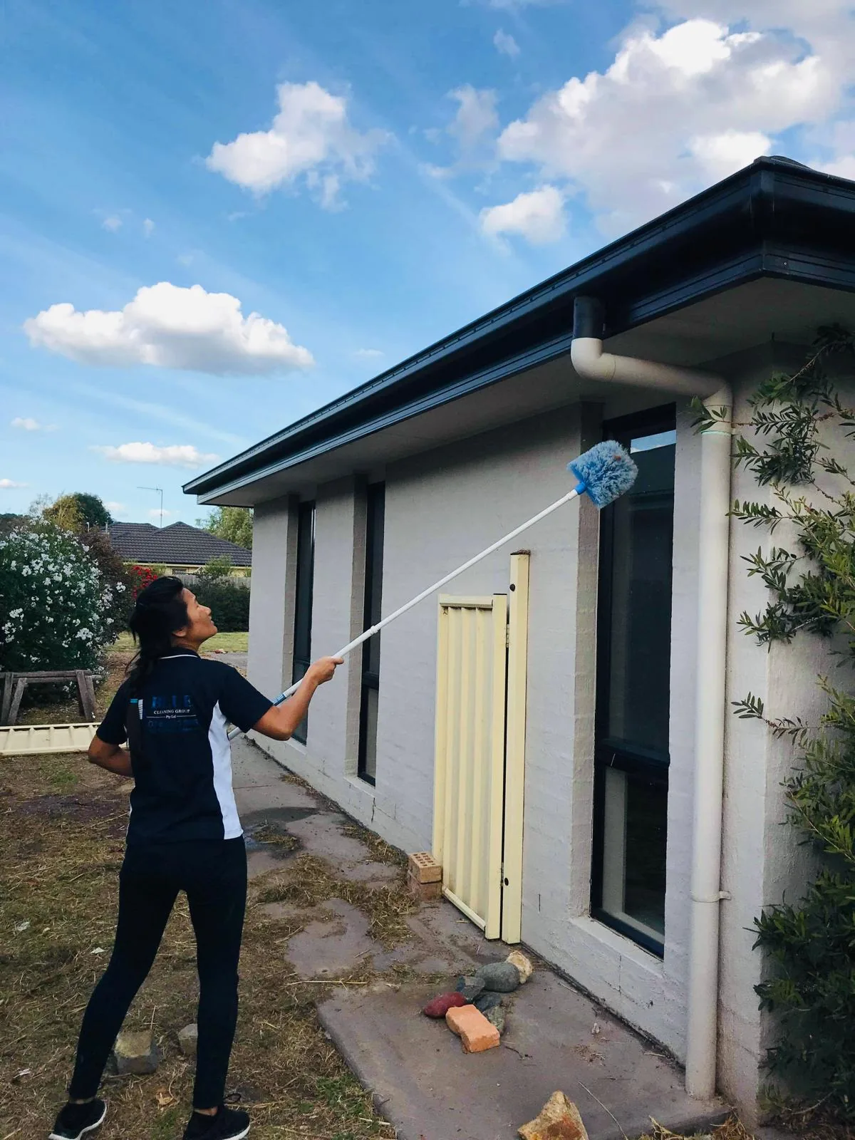 A Blue Cleaning Group cleaner raising a cobweb duster on an extension pole to the eaves of a rendered house