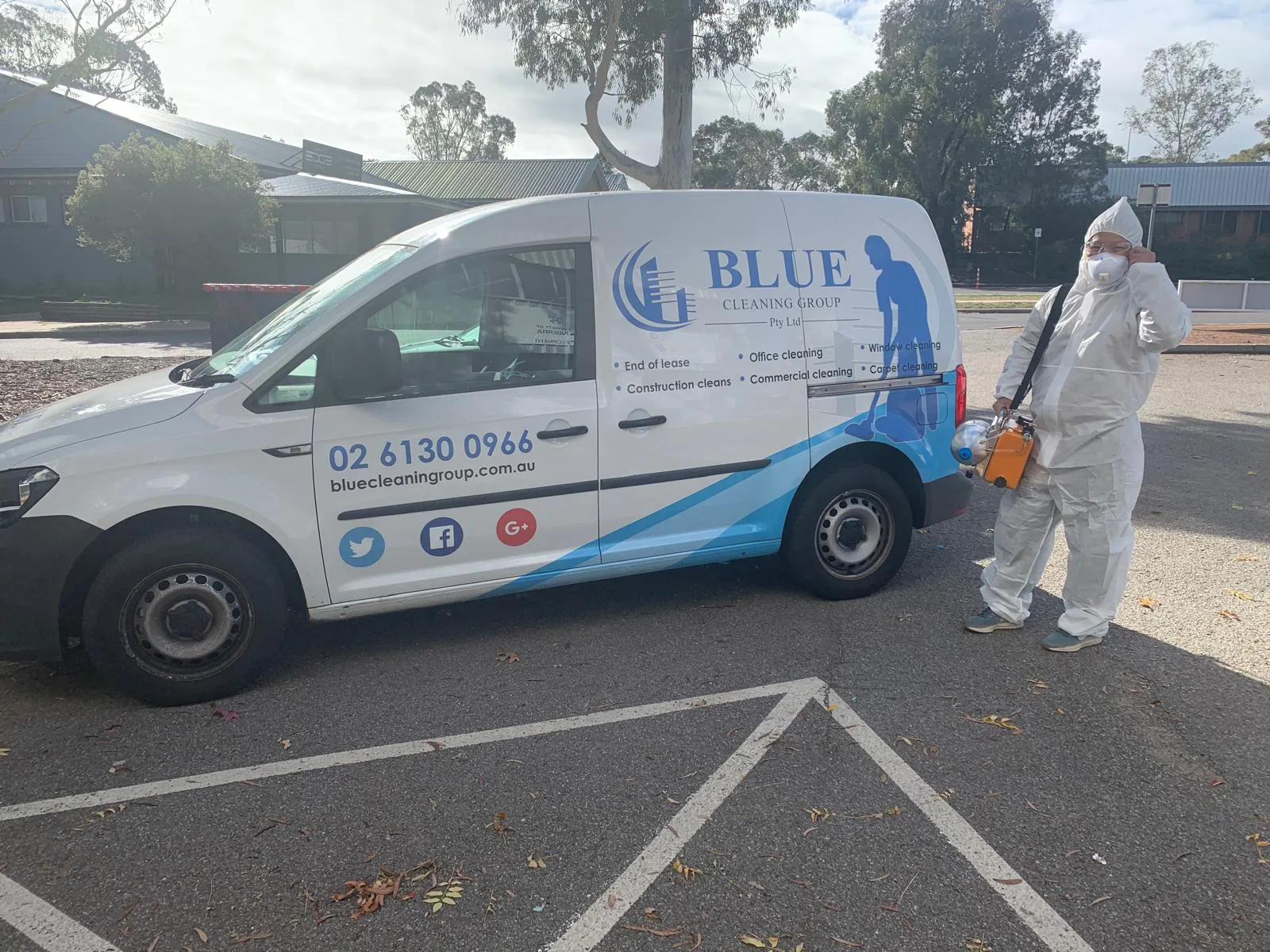 A worker in a full white disposable coverall and mask with a fogging machine, beside the Blue Cleaning Group van