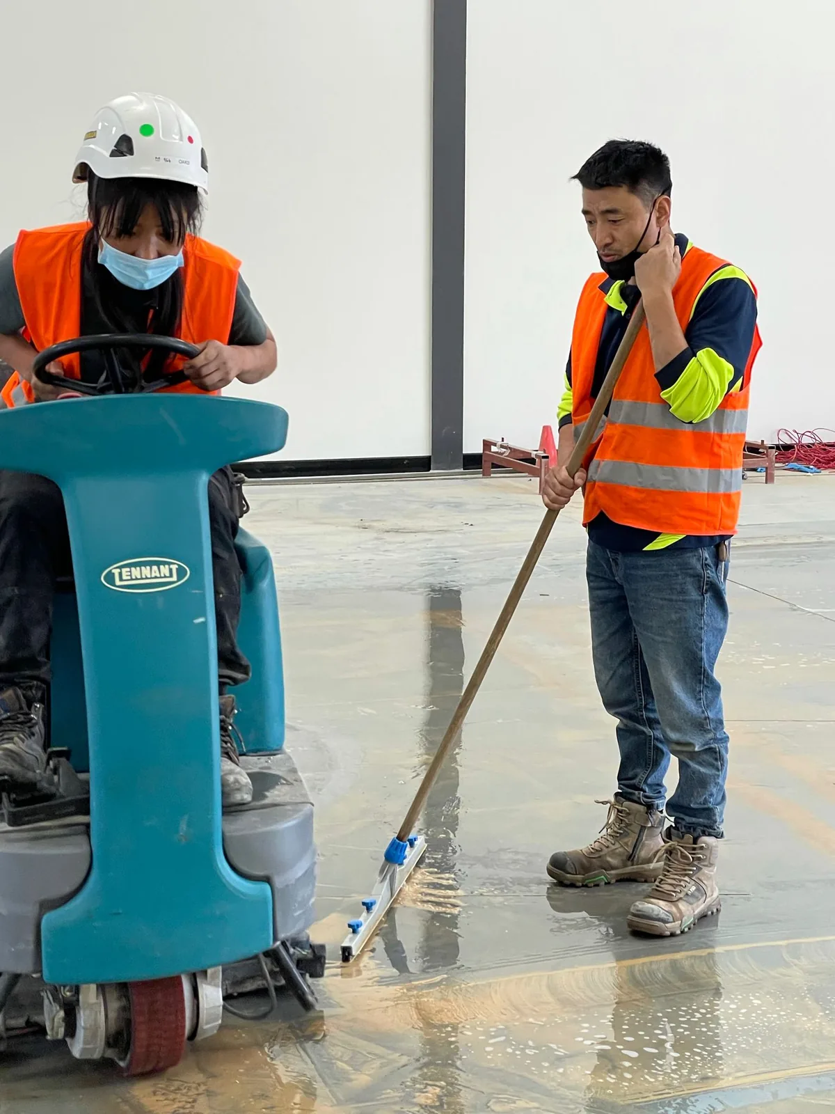 One worker on a Tennant ride-on scrubber while another squeegees the wet concrete floor of an empty commercial shell
