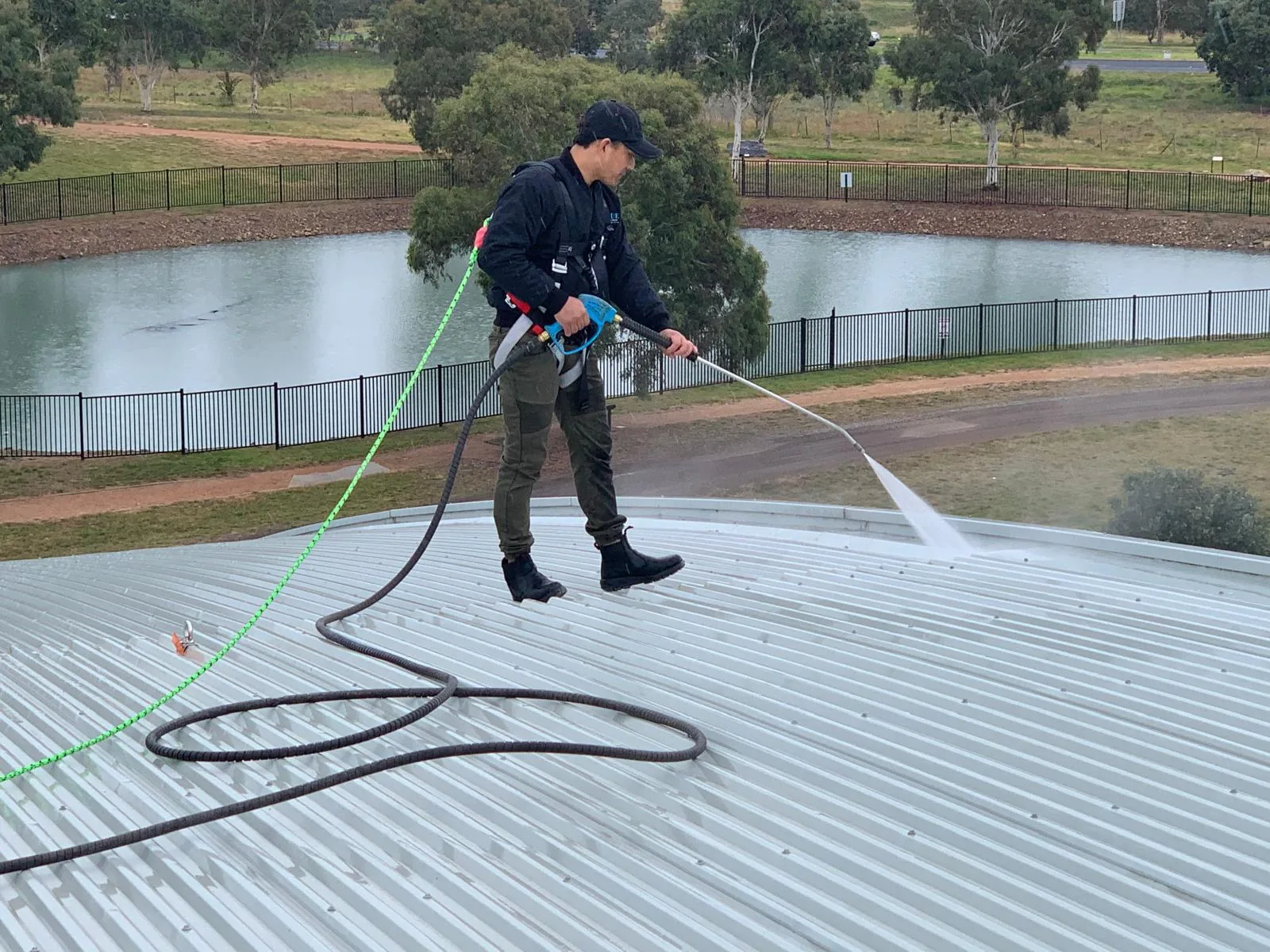 A harnessed worker pressure washing a corrugated metal roof, hose and lifeline trailing across the sheeting