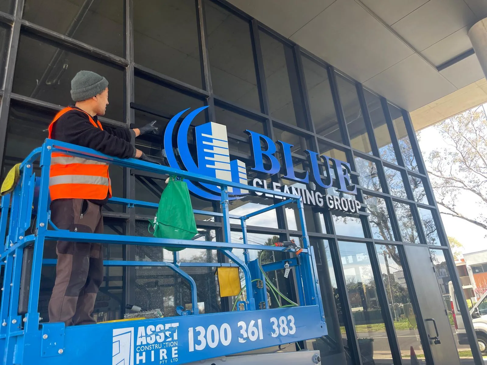 A worker on a scissor lift wiping glazing beside the Blue Cleaning Group sign on a glass facade
