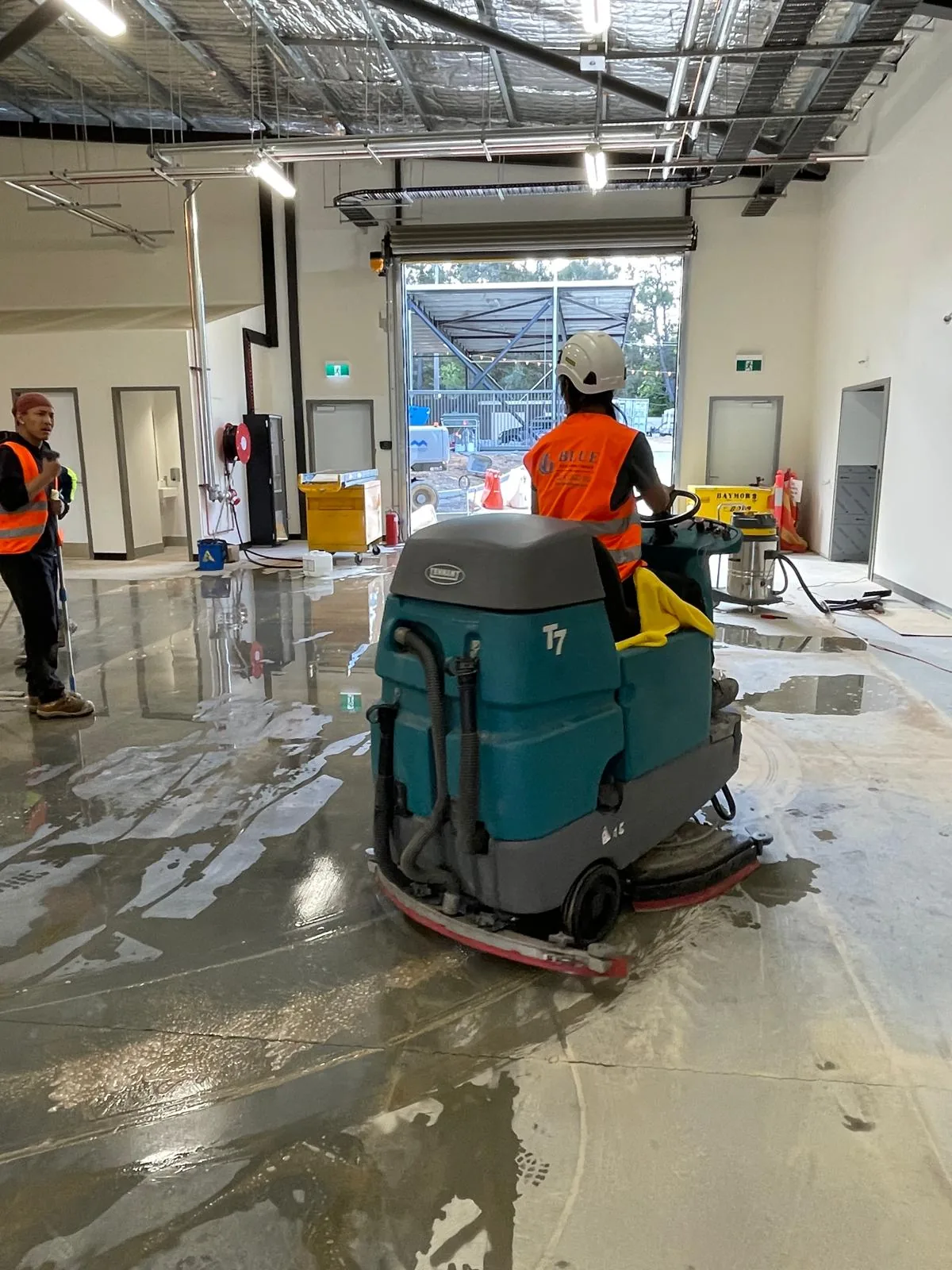 An operator in a Blue Cleaning Group vest driving a Tennant T7 ride-on scrubber over a wet slab towards an open roller door