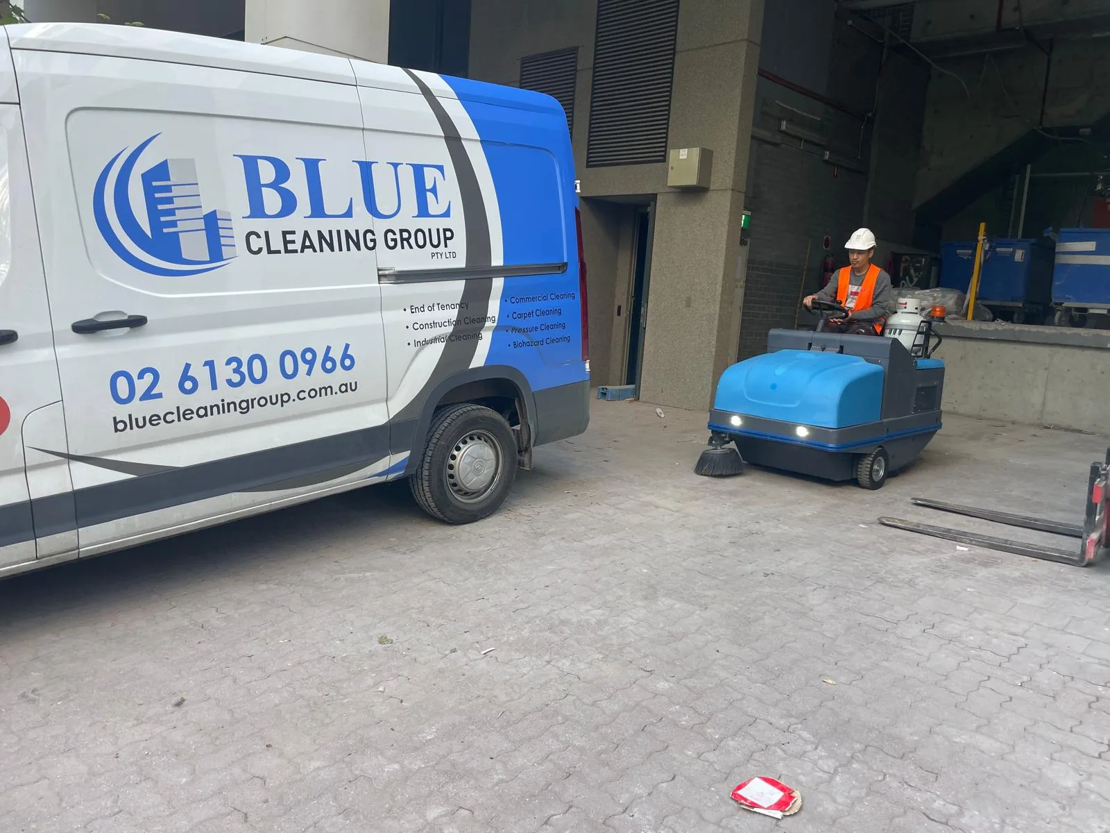 The signwritten Blue Cleaning Group van on a loading bay while a worker drives a ride-on sweeper across the pavers