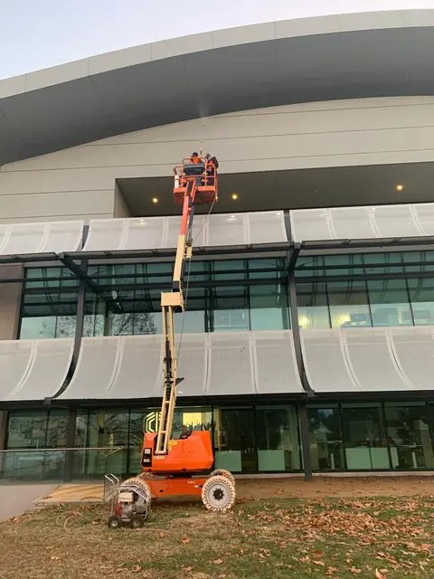 Two workers in a raised boom lift basket washing down the curved metal fascia of a commercial building