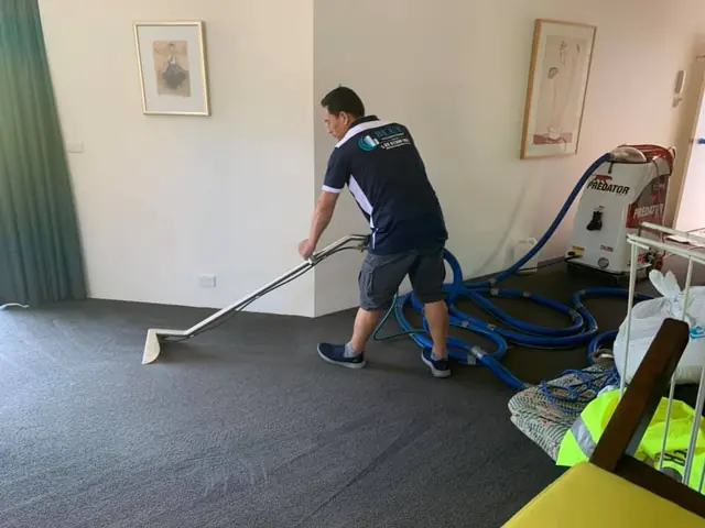 A Blue Cleaning Group operator drawing a carpet extraction wand across the carpet of a furnished lounge