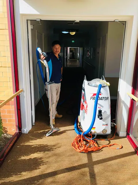 A cleaner standing in a corridor doorway with a carpet extraction wand beside a portable extractor