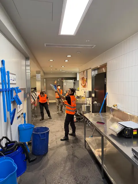 Three workers in orange Blue Cleaning Group vests working a narrow commercial kitchen galley, one reaching a mop to the ceiling