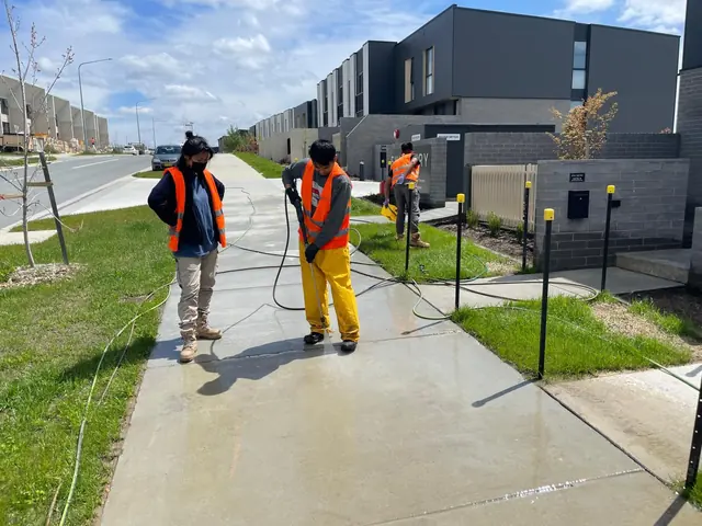 Three workers in orange hi-vis pressure washing the concrete footpath outside newly built townhouses