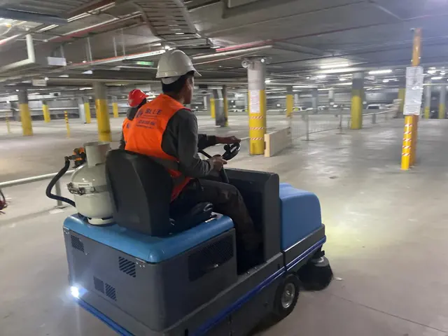 A worker in a hard hat driving an LPG ride-on sweeper across an empty multi-storey car park deck