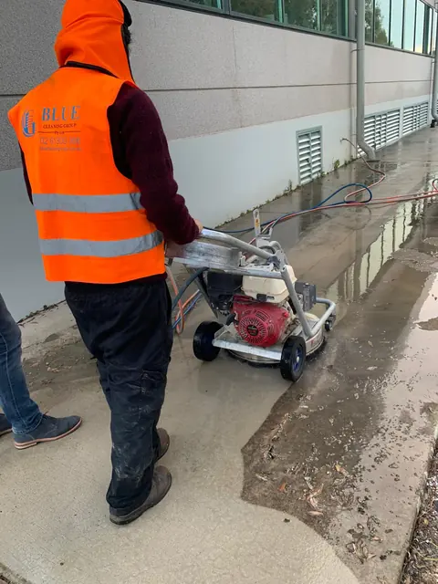 A worker in a Blue Cleaning Group hi-vis vest pushing a rotary surface cleaner along a wet footpath