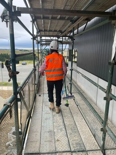 A worker in a Blue Cleaning Group hi-vis vest walking a scaffold walkway past dark cladding, brooms on the boards