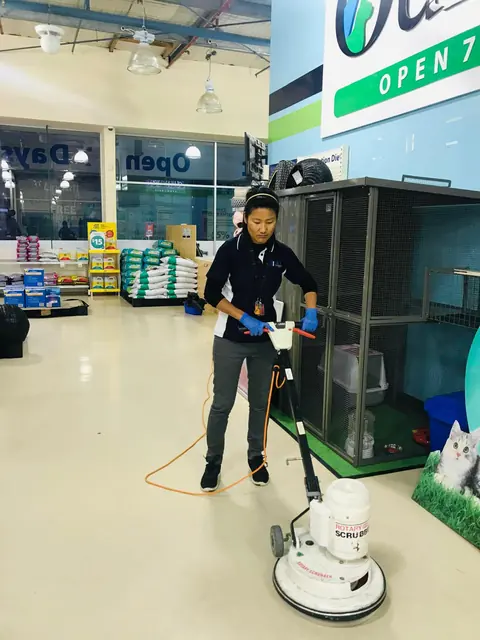 A cleaner in a navy Blue Cleaning Group polo running a rotary scrubber across the vinyl floor of a pet supplies shop
