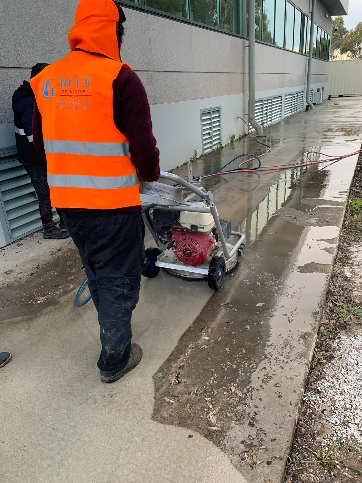 A worker in a printed Blue Cleaning Group vest running a rotary surface cleaner along a wet footpath