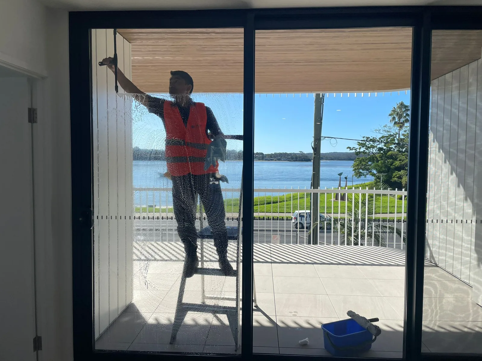 A cleaner on a ladder cleaning a balcony window with water visible beyond