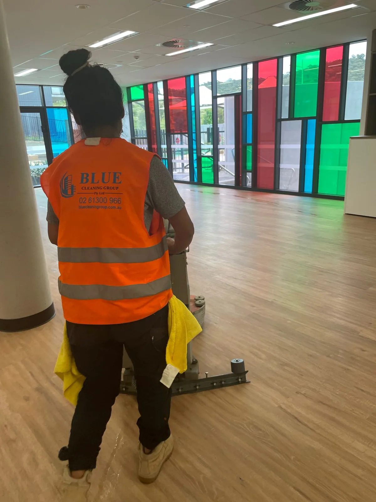 A worker in a Blue Cleaning Group vest running a rotary floor machine through a hall glazed with coloured panels